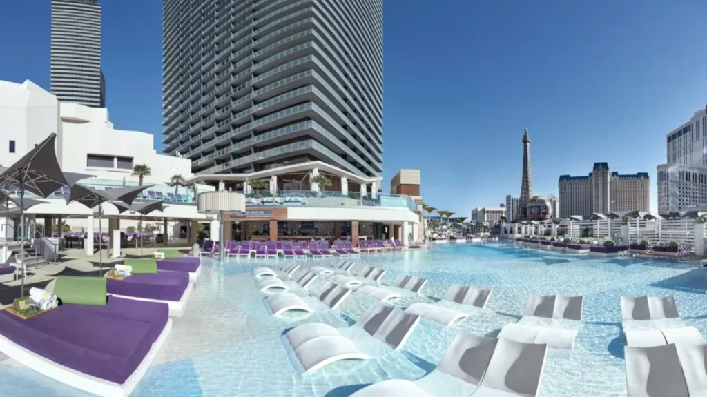 Panoramic view of the main deck of the Cosmopolitan Pool District, showing submerged daybeds and the Las Vegas Strip skyline in the background.